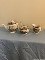 Teapot, sugar bowl with lid, and creamer displayed together on table, showing friendly village pattern in multiple colors.