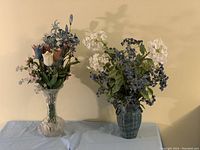 Wide view of both floral arrangements on a table under neutral wall shown to capture the overall appearance and condition.