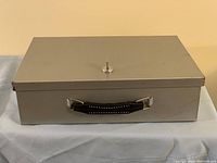 Closed metal fire safe box with a key inserted in the lock and a black handle on the front side.