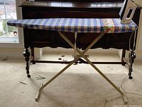 Full view of ironing board with blue and white checkered strawberry fabric cover and iron standing on end