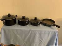 Set of three black Lagostina pots with glass lids and one frying pan arranged on a table with blue cloth, showing full set from side.