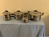 Wide shot showing all four KitchenAid stainless steel pots with lids on a light blue cloth.