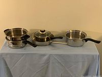 Full set of frying pan, two pots, and double boiler displayed on table with a light blue cloth.