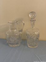 Decanter with stopper and matching pitcher showing pinwheel cut pattern on clear crystal, set on neutral background.