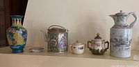 Five items displayed in a row: metal vase, Chinese teapot, small sugar pot, porcelain teapot, and ceramic jug.