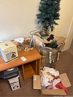 View showing two Christmas trees including a tall tree in metal urn base and smaller snow-flocked tree, a boxed snow globe, and ceramic holiday village buildings on a wooden desk plus a cardboard box with packing material and decorations