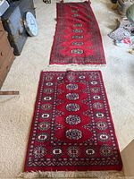Two red hand pulled rugs displayed lengthwise on beige carpet, showing geometric patterns and fringed edges.