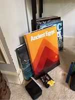 Photo showing five history books with visible titles 'Ancient Egypt' and 'The Age of Expansion' and a set of binoculars with case and cleaning cloth on floor.