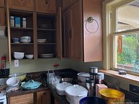 Kitchen countertop with various ceramic casserole dishes, bowls, pans, and kitchen essentials near wooden cabinets and window.