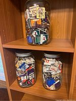 Three large glass jars displayed on a wooden shelf filled with assorted vintage match boxes and match books of various brands and designs.