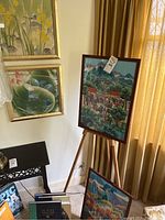 Two framed artworks displayed on wooden tripod easel with two additional framed pieces visible on wall and floor