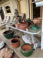 Table filled with terracotta pots, green glazed pots, stacked saucers, watering can, and decorative statue near outdoor chairs