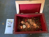 Wooden box with red felt lining filled with pennies. A variety of copper pennies can be seen, some showing wear and discoloration. Box clasp is broken, visible on top edge of box.