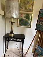 Photo of black side table with decorative apron and brass lamp with cream fabric shade placed on top against corner wall with paintings.