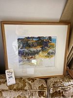 Framed and matted watercolor painting showing a person with a hat sitting outdoors facing a lake or body of water surrounded by trees and grass.