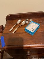 Two sterling silver spoons, a ring in a blue box, and salt and pepper shakers with sterling silver tops placed on a wood surface.