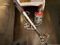 Dyson vacuum cleaner unit with hose and wand attachments shown on floor next to couch and table.