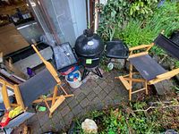 Wide overview showing black ceramic egg grill on metal tripod stand with folding side shelf, two folding director chairs, charcoal bag, tool case, and ceramic crock