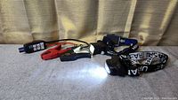 Two black LED headlamps on adjustable elastic straps and quick start battery booster pack with clamps and cables on grey surface, one headlamp lit.