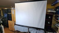 Front view of the large white projector screen partially extended, mounted on a tripod stand in a cluttered room.