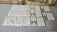 Photo showing 3 Apple wired keyboards with white keys and aluminum casing and 2 Belkin wireless numeric keypads arranged on a gray surface. USB cables are visible on keyboards.