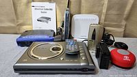 Photo showing the lot items arranged in a row including 3 routers (Linksys, Netgear, SmartRG), portable DVD player, personal tape recorder, USB power block, Bluetooth speaker, and remotes.