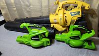 Lot showing three yard tools: yellow Yard-Man gas-powered leaf blower and two green Greenworks electric leaf blowers on a grey background.