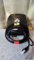 Closed black Mickey Mouse waffle maker with red latch and power indicator. Shows front design with Mickey Mouse character.