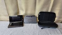 Nintendo 3DS handheld console open next to black docking station and black carrying case on fabric surface