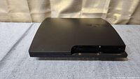 Top front angle view of the black PlayStation 3 Slim console showing the disc slot and partial surface details.