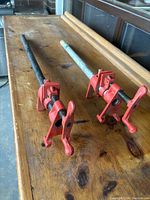 Two red metal pipe clamps positioned on a wooden surface, displaying the full length of the pipe and red clamp mechanism.