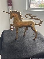 Photo showing the left profile of the solid brass unicorn figurine standing on a black cloth with a window and building in the background.