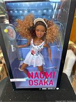 Clear front view of the Naomi Osaka Barbie doll in original packaging, showing detailed doll with tennis outfit and accessories.