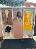 Photo of Barbie doll and multiple wardrobe items including a brown coat, yellow dress, black shorts, and shoes in original box.