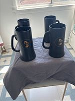 Four matt black glass mugs arranged on a cloth, showing the exterior handles and gold insignia and figures on the sides.