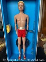 Front view of doll in blue packaging with two interchangeable hands and red swim trunks, showing articulated arms and legs.