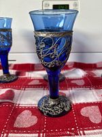 Tall blue glass with ornate filigree pewter wrapping on bowl and round base, intricate curling patterns, on a red and white heart patterned cloth.