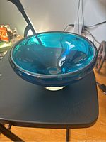 Full view of a deep vintage heavy blue glass bowl placed on a black surface, showing its rounded shape, clear thick base, and visible air bubbles embedded in the blue glass.