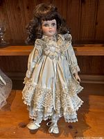 Front view of vintage porcelain girl doll standing on wooden surface, wearing cream lace pinafore dress and matching shoes.