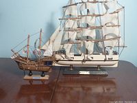 Two wooden model sailing ships side by side on a wooden surface against a pale blue wall, showing side profile and height comparison.