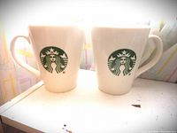 Two white ceramic Starbucks mugs front-facing showing the green siren logo each with handles to the right.