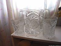 Clear glass pitcher with two pressed glass tumblers featuring a detailed pinwheel pattern arranged on a white surface in front of a window with light curtains.