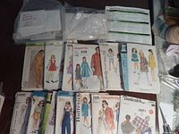 Sewing pattern envelopes arranged on wooden surface showing various dresses, jumpsuits, and outfits for women and children. Includes Simplicity, Butterick, and other vintage sewing pattern brands.