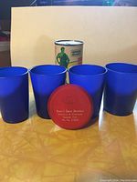 Four blue stacking cups with one red lid showing 'Ted's Esso Service' imprint, and Green Giant tin in background