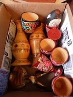 Box containing various wooden items including painted wooden egg cups, carved wooden vases with etched patterns, and a black and red painted wooden egg.