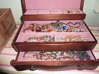 Opened 3-tier jewelry box showing pink felt lining with multiple compartments containing assorted costume jewelry including necklaces, bracelets, rings, and earrings.