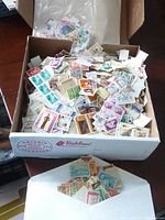 Large cardboard box filled with a variety of loose postage stamps of different colors and designs.