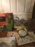 Multiple pieces of artwork leaning on a surface, including paintings of houses and trees, botanical and floral prints.