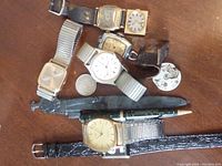 Top view of six men's wristwatches laid out with a carved black figure and green pen, showing various watch styles and bands.