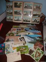 Photo showing the open postcard album with multiple postcards in sleeves featuring buildings and scenic views, along with a collection of loose postcards spread on a table.